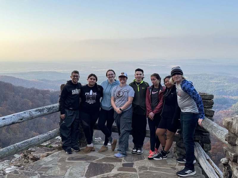 A group of eight people stands smiling on a stone lookout point overlooking a vast forested valley. The mood is cheerful and the landscape is serene.