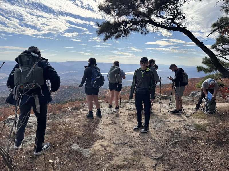 A group of hikers with backpacks stands on a rocky overlook, enjoying a scenic view of mountains and an expansive valley under a partly cloudy sky.