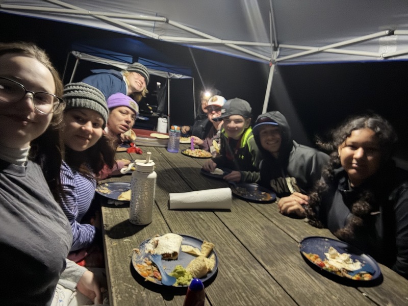 A group of people sits around a picnic table under a canopy at night, sharing a meal. They seem cheerful and cozy, wearing hats and jackets.