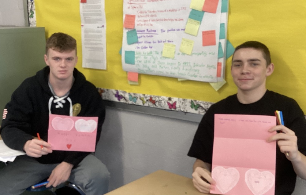 Two people hold pink Valentine's cards with heart designs, sitting in a classroom. A yellow bulletin board with notes is in the background.