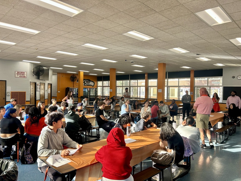 Students listening to man speak in cafeteria.