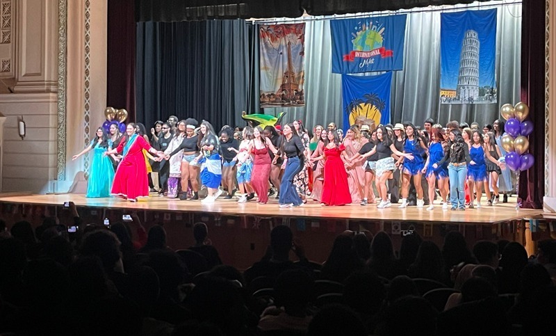 A large group of people in colorful outfits perform energetically on stage, under banners of iconic landmarks, conveying excitement and celebration.