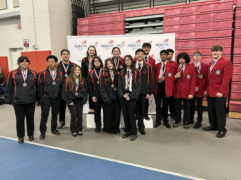 A group of students wearing medals stands in front of a "SkillsUSA" banner. They're dressed in black and red jackets or red blazers, smiling proudly.