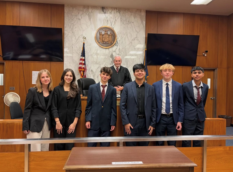 A group of six young people in formal attire stand smiling in a courtroom, with an older male judge behind them. The setting is formal and professional.