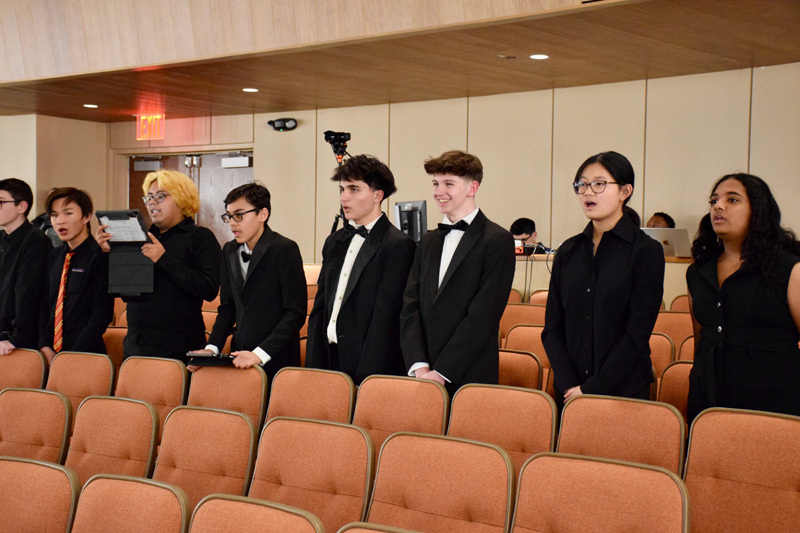 A group of seven young adults, dressed in formal black attire, stand in a theater's empty seating area