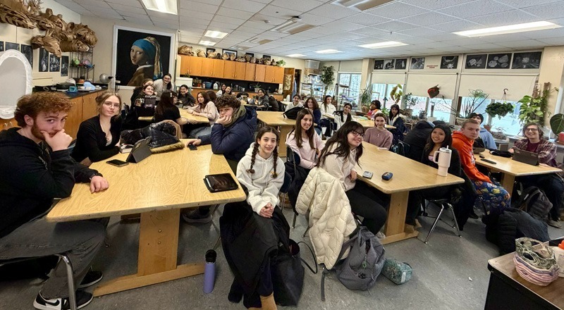 A classroom filled with students sitting at wooden tables, attentively facing forward. The room is decorated with art and plants, creating a lively atmosphere.