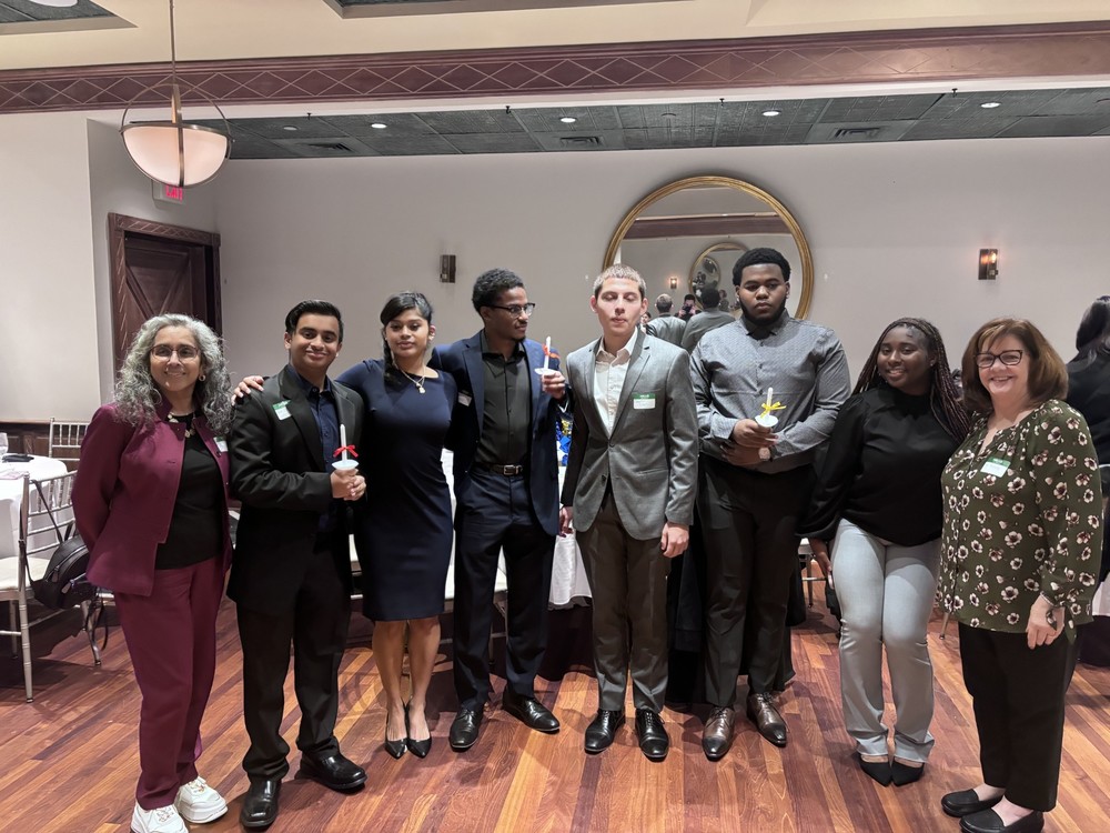 A group of eight people stands in formal attire in a well-lit room with wooden floors.