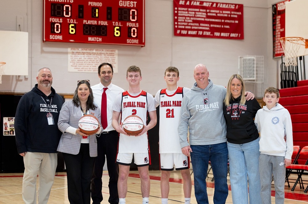 A group stands on a basketball court under a scoreboard. Two players hold basketballs, surrounded by coaches and family, smiling proudly.