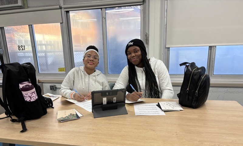 people sit at a table, smiling and writing on paper near a tablet. 