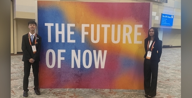 Two young individuals stand beside a vibrant banner displaying the text 'THE FUTURE OF NOW' at a conference event.