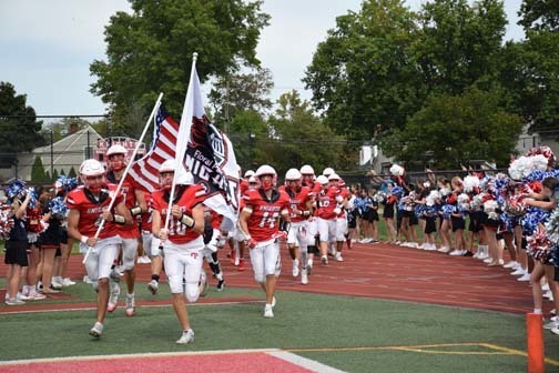 Football team running with flags, with cheerleaders on the sides.