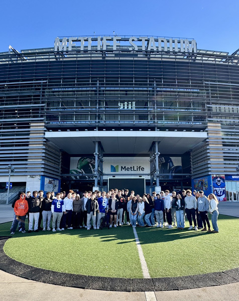 A large group of people stands in front of MetLife Stadium, surrounded by a clear blue sky and bright sunlight.