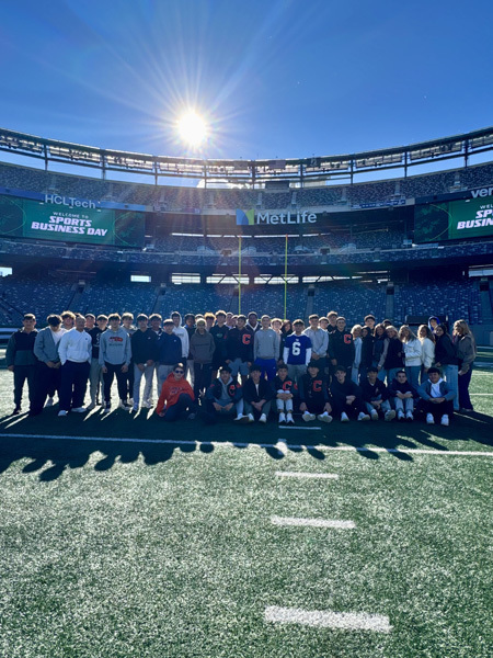 A large group of people stands in front of MetLife Stadium, surrounded by a clear blue sky and bright sunlight.