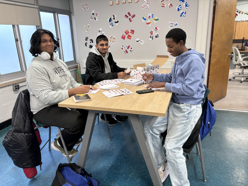 people sit at a table, smiling and writing on paper near a tablet. 