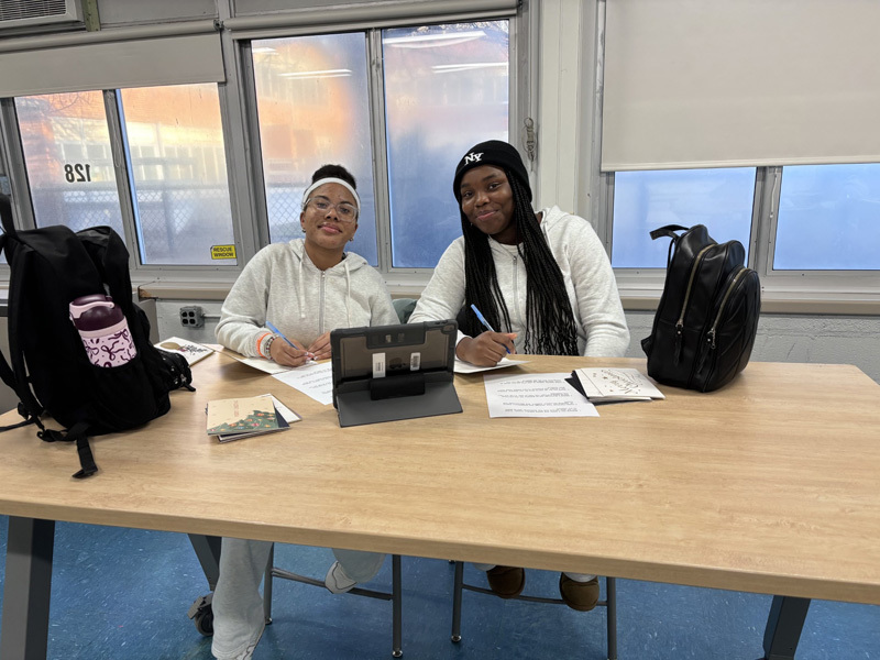 people sit at a table, smiling and writing on paper near a tablet. 