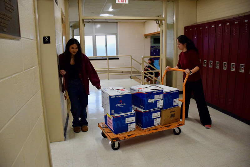  group of young adults pose together in a school hallway, with a  cart with boxes of items