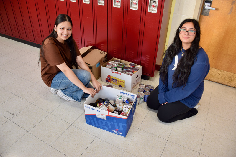  group of young adults pose together in a school hallway, smiling. with donations in boxes on floor