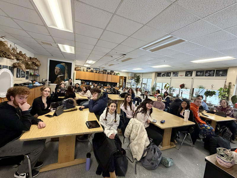 A classroom filled with students sitting at wooden tables, attentively facing forward. The room is decorated with art and plants, creating a lively atmosphere.