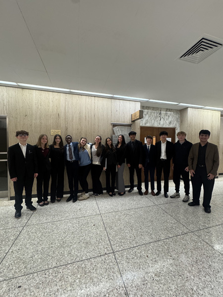 A group of eleven people stands in a well-lit hallway with neutral walls and tile flooring, dressed in formal attire, smiling and relaxed.