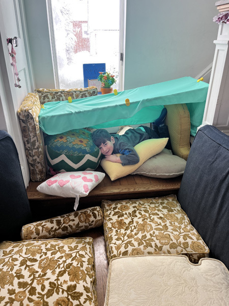 A child smiles while lying in a cushion fort with turquoise fabric and patterned pillows. The cozy setup is near a window, creating a playful scene.