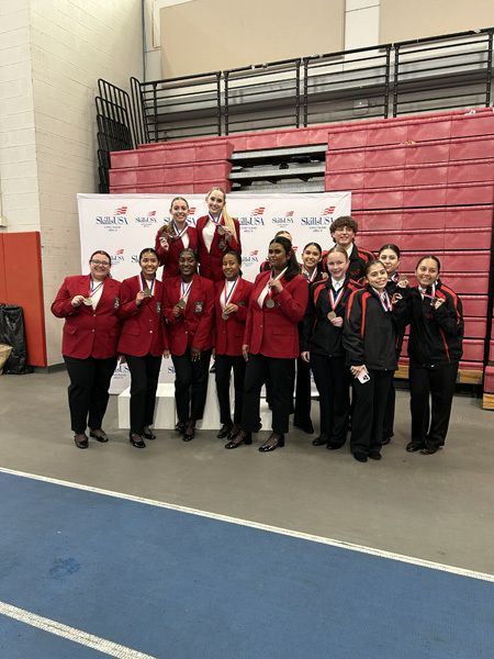 Three young women in red blazers with patches stand smiling in front of a SkillsUSA Long Island Area VI backdrop, exuding pride and confidence.