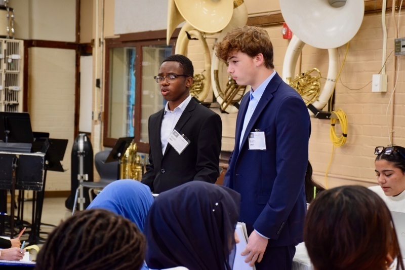Presenter speaking to an engaged audience in a classroom setting during a conference or workshop, with students seated and taking notes.