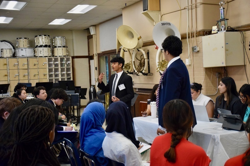 Presenter speaking to an engaged audience in a classroom setting during a conference or workshop, with students seated and taking notes.