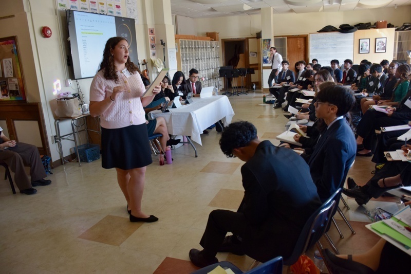 Presenter speaking to an engaged audience in a classroom setting during a conference or workshop, with students seated and taking notes.