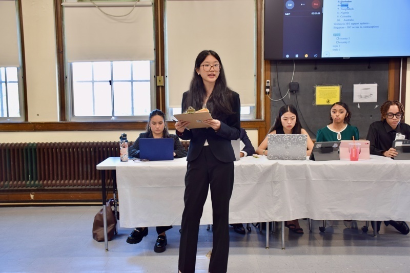 Presenter speaking to an engaged audience in a classroom setting during a conference or workshop, with students seated and taking notes.