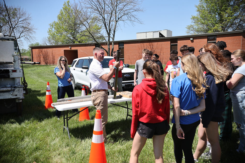 KDOT geologist holds a core sample while speaking to a group of 8th grade students gathered outdoors near a drill rig.