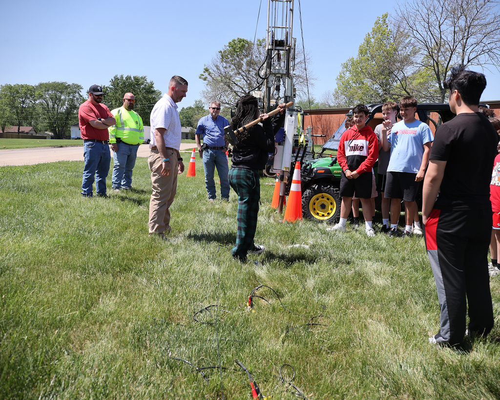 Student participates in a hands-on geology activity using field equipment as classmates observe and smile.
