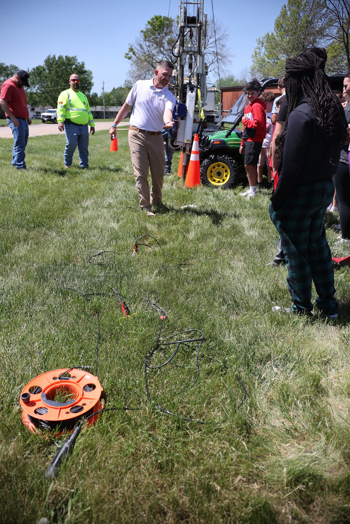 KDOT staff member demonstrates seismic testing equipment laid out on the grass while students watch nearby.