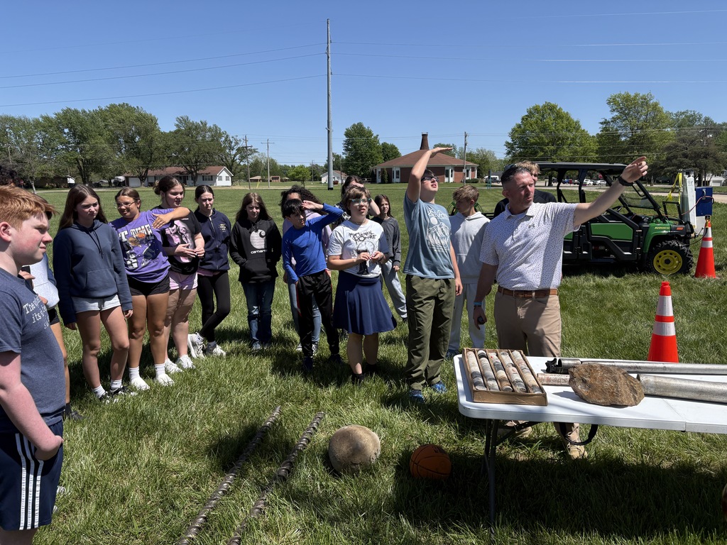 KDOT geologist points into the distance while explaining rock samples and drilling tools displayed on a table to students.