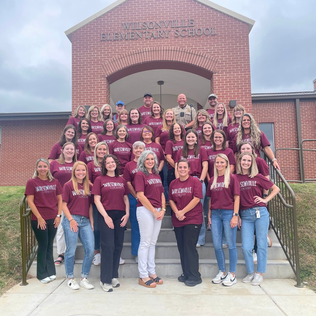 faculty and staff standing in an organized fashion in front of the school's entrance all wearing the same shirt saying Wilsonville Elementary