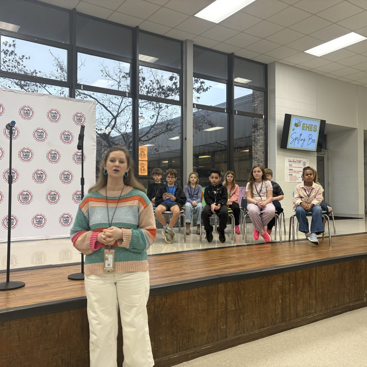 spelling bee coordinator speaking and participants sitting on the stage behind her 
