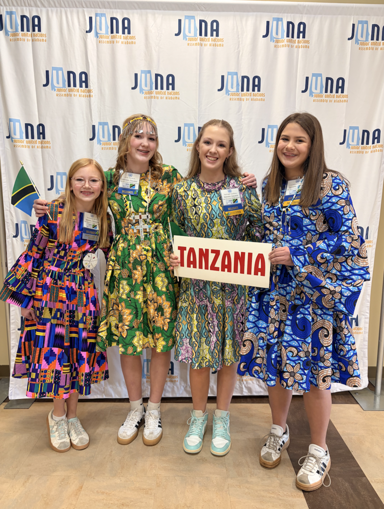 JUNA Tanzania Delegation holding their "Tanzania" placard in front a JUNA photo backdrop, wearing their brightly colored costumes.