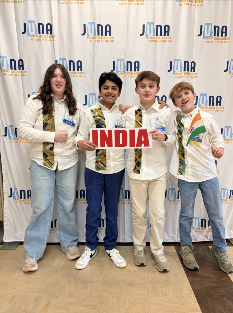 JUNA India Delegation holding their "India" placard in front a JUNA photo backdrop, wearing their costumes.