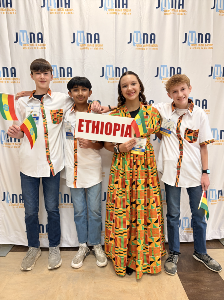 JUNA Ethiopia Delegation holding their "Ethiopia" placard in front a JUNA photo backdrop, wearing their costumes.