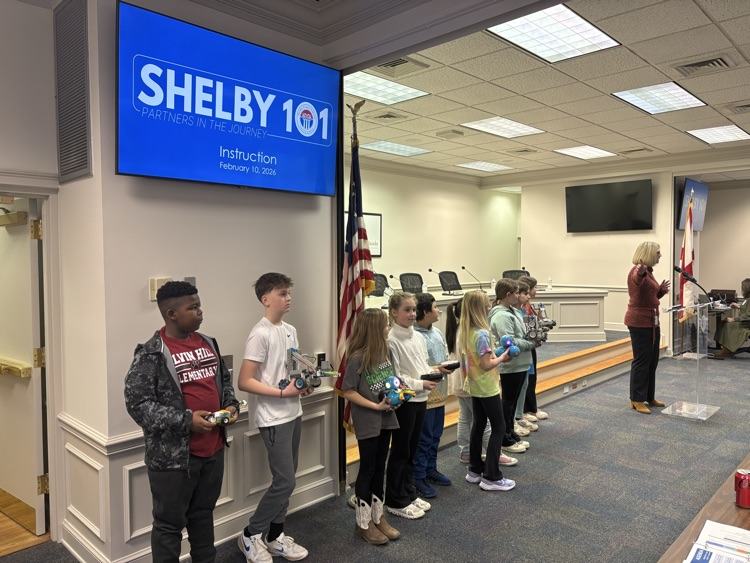 District representative speaks while students hold robots and coding devices