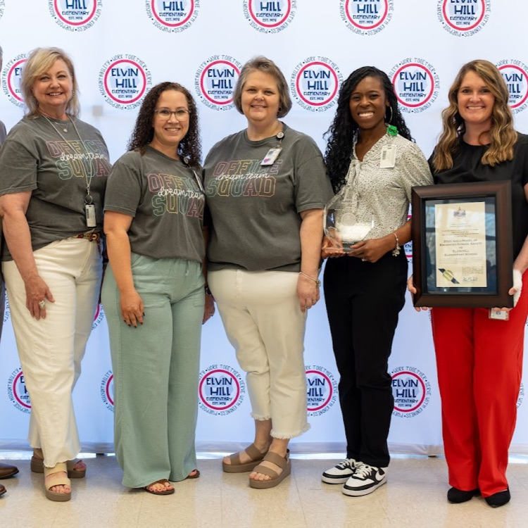 5 ladies in front of school backdrop holding a framed certificate and acrylic trophy