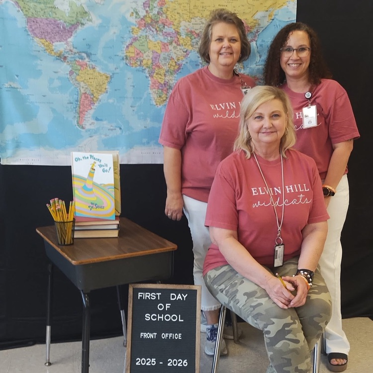 one woman seated at a desk with 2 women standing behind her in front of a backdrop with a map and book cover 