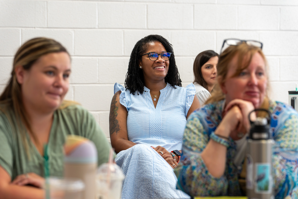An educator smiles while listening during a professional learning session with fellow teachers seated around the room.
