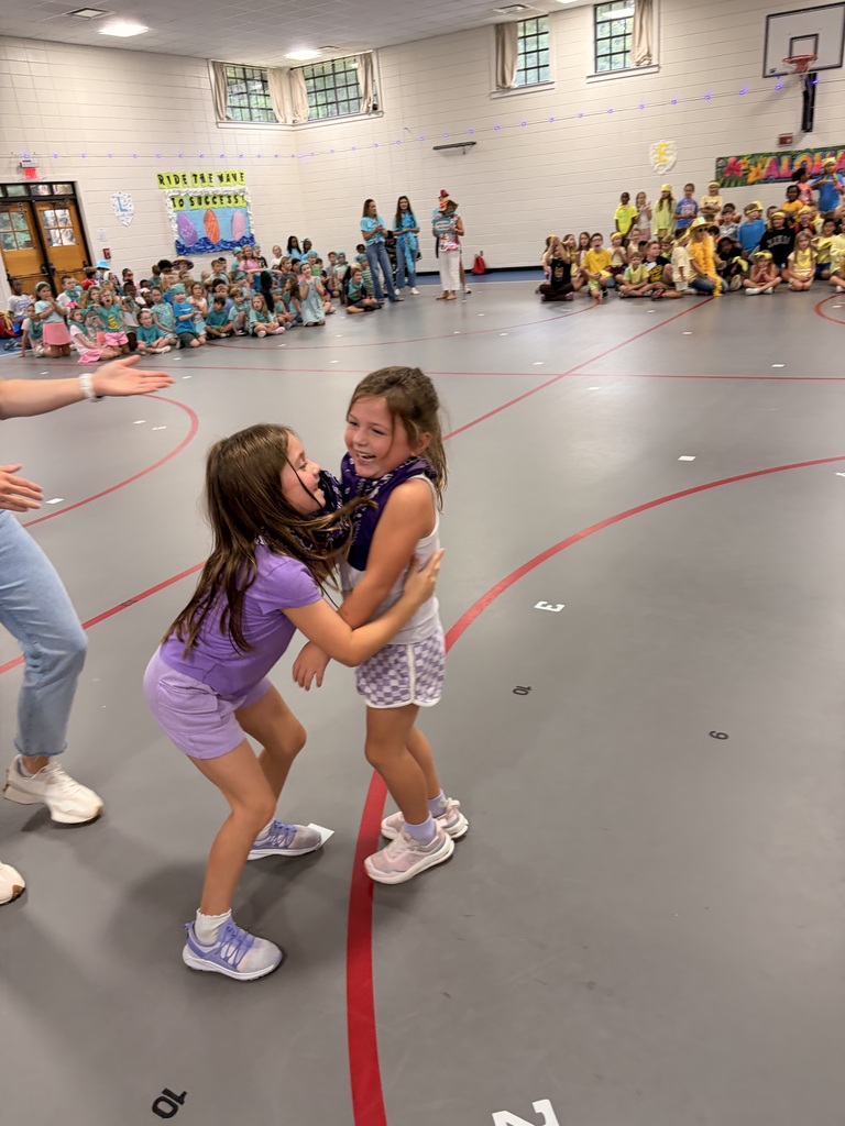 Two girls wearing purple bandanas hugging, with other groups of students gathered in the background.