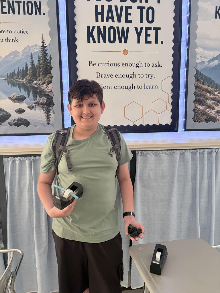 A middle school student poses in front of shelves in his math classroom. he's wearing a green shirt and is proudly holding a tape dispenser that he has repaired in his right hand, and the parts he 3d printed in his left hand.  