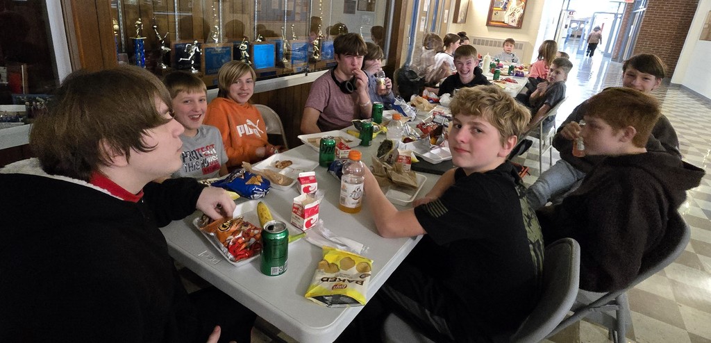 Junior high students sitting at tables in the main hallway as the staff serves them for their pizza party