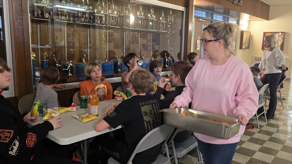 Junior high students sitting at tables in the main hallway as the staff serves them for their pizza party
