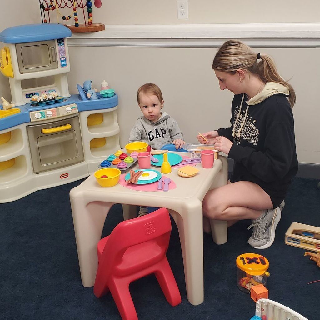A high school student playing with a young toddler at a small table with plastic food and dishes