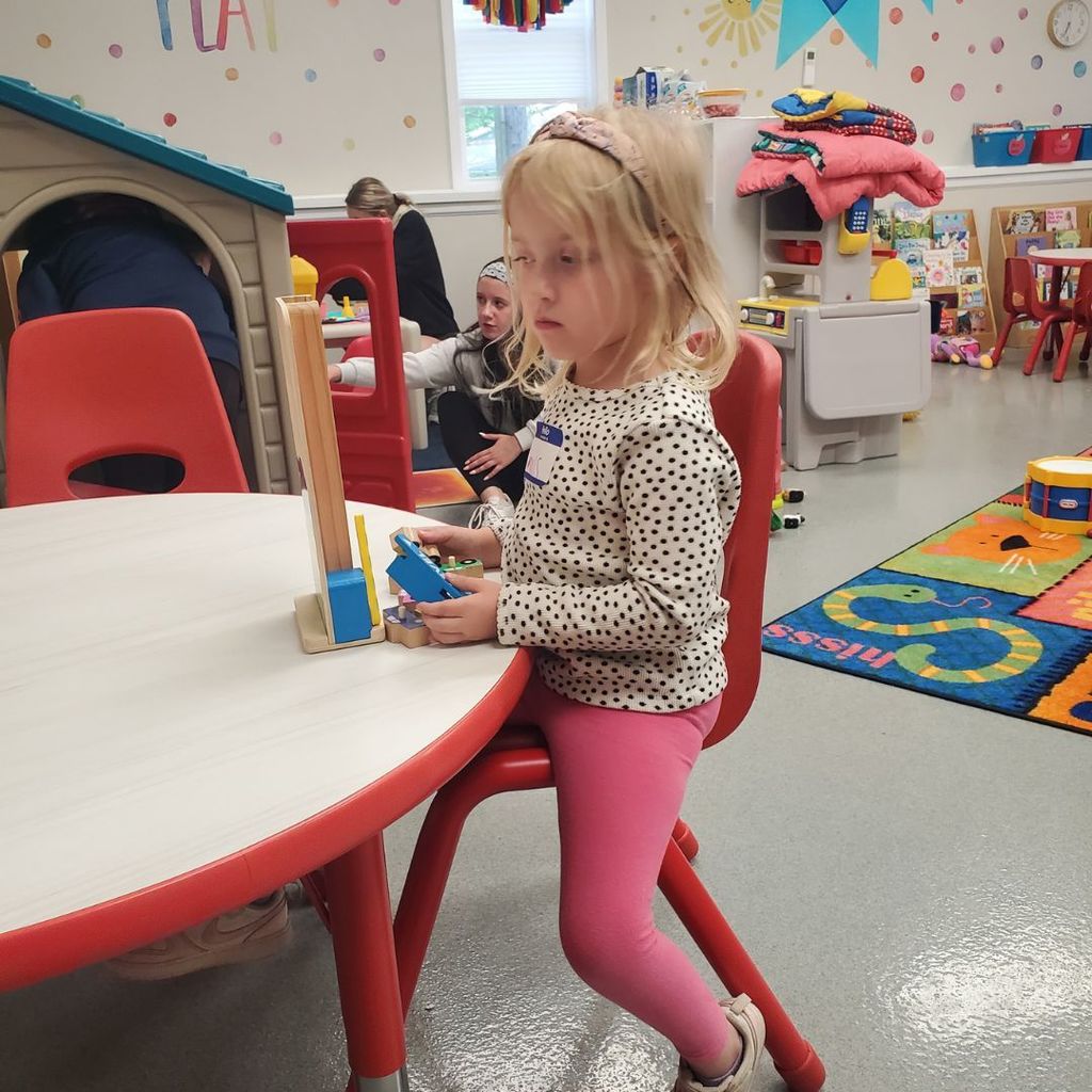 Photo of young girl playing at a table while other children can be seen playing in the background