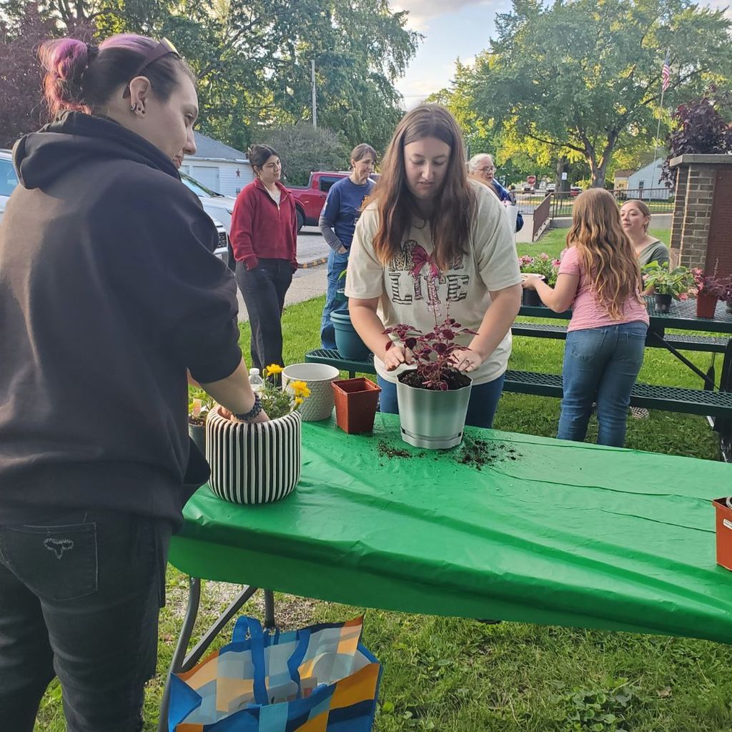 Two women putting flowers in pots of dirt at an outside table while others can be seen with flowers at a table in the background