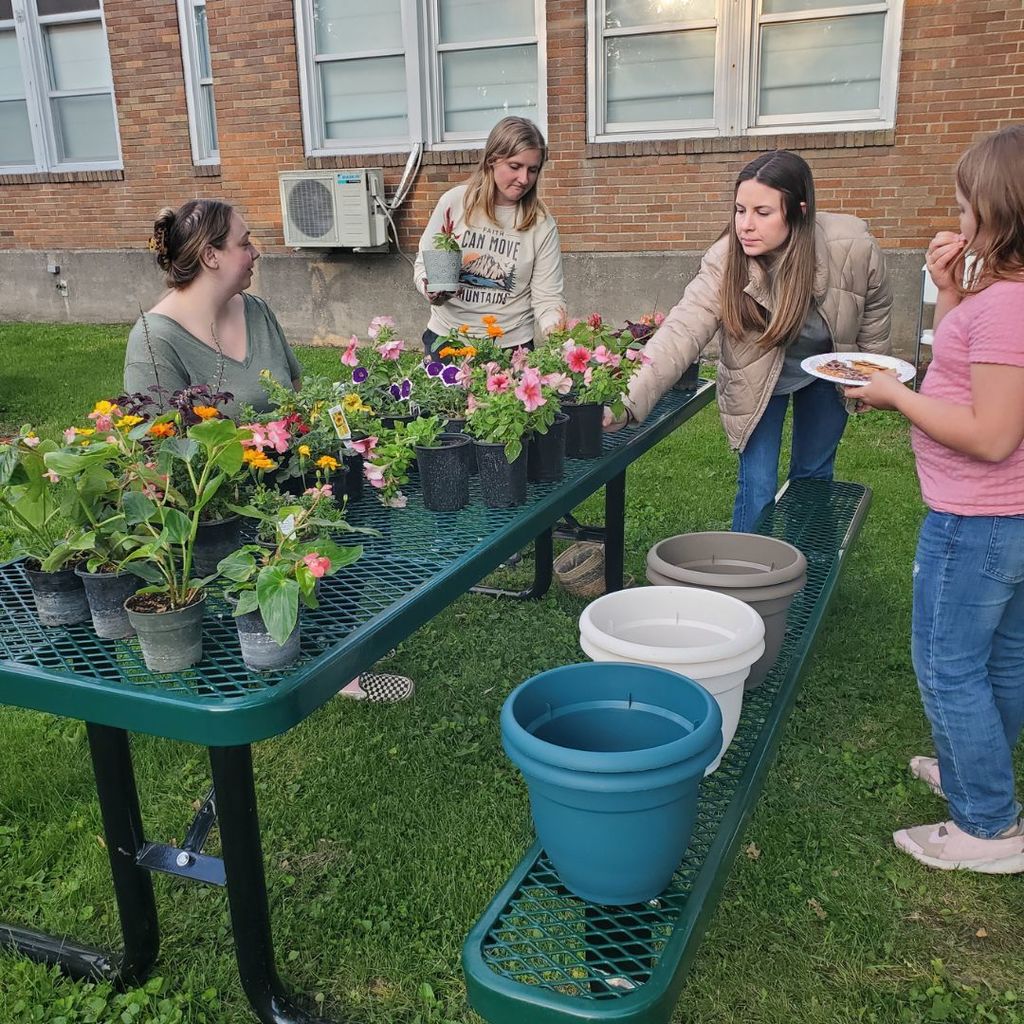 Three woman examining flowers on an outside table while a young lady stands to the right and looks on